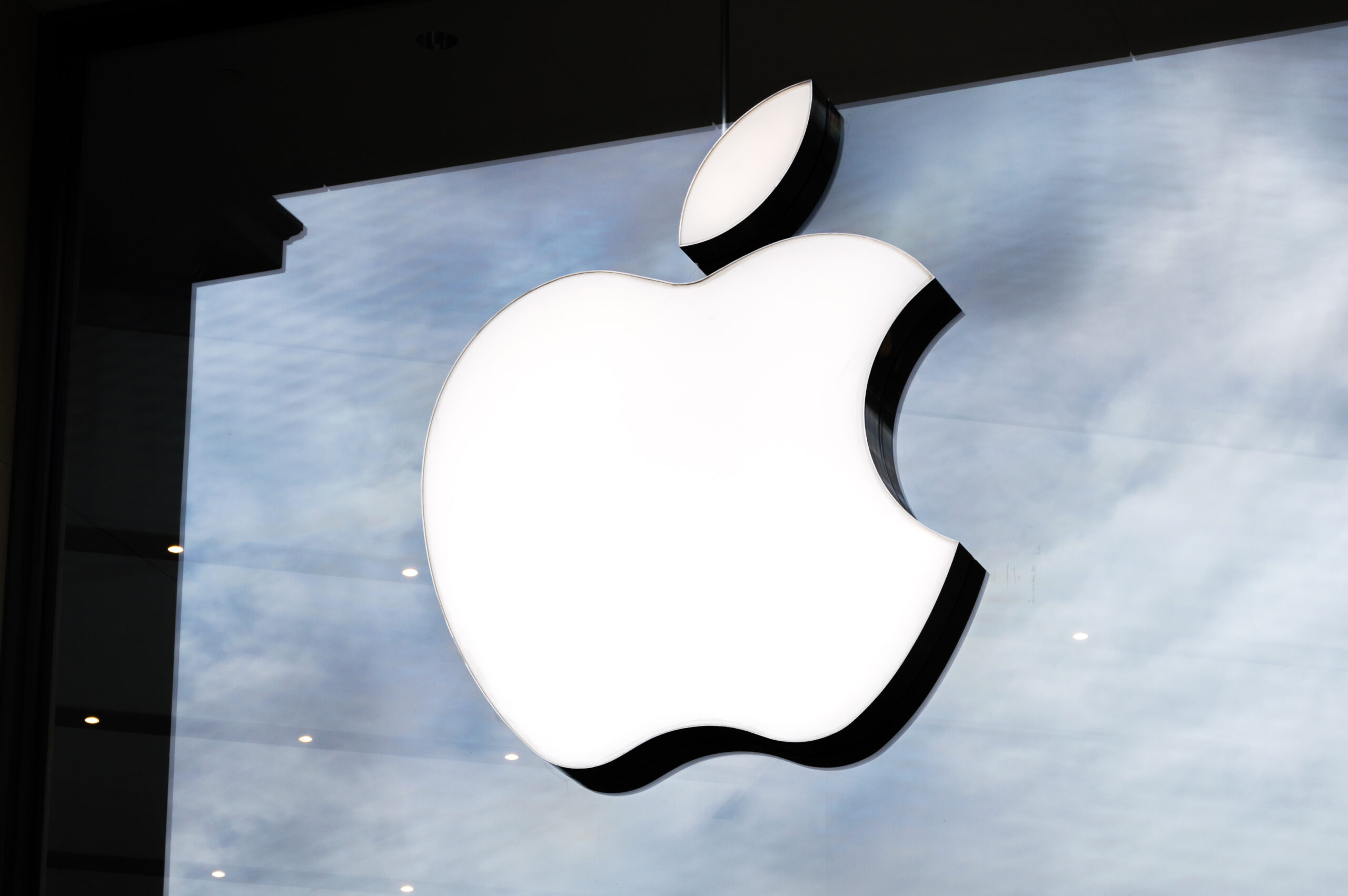 Illuminated Apple logo on a glass storefront with a cloudy sky reflection in the background