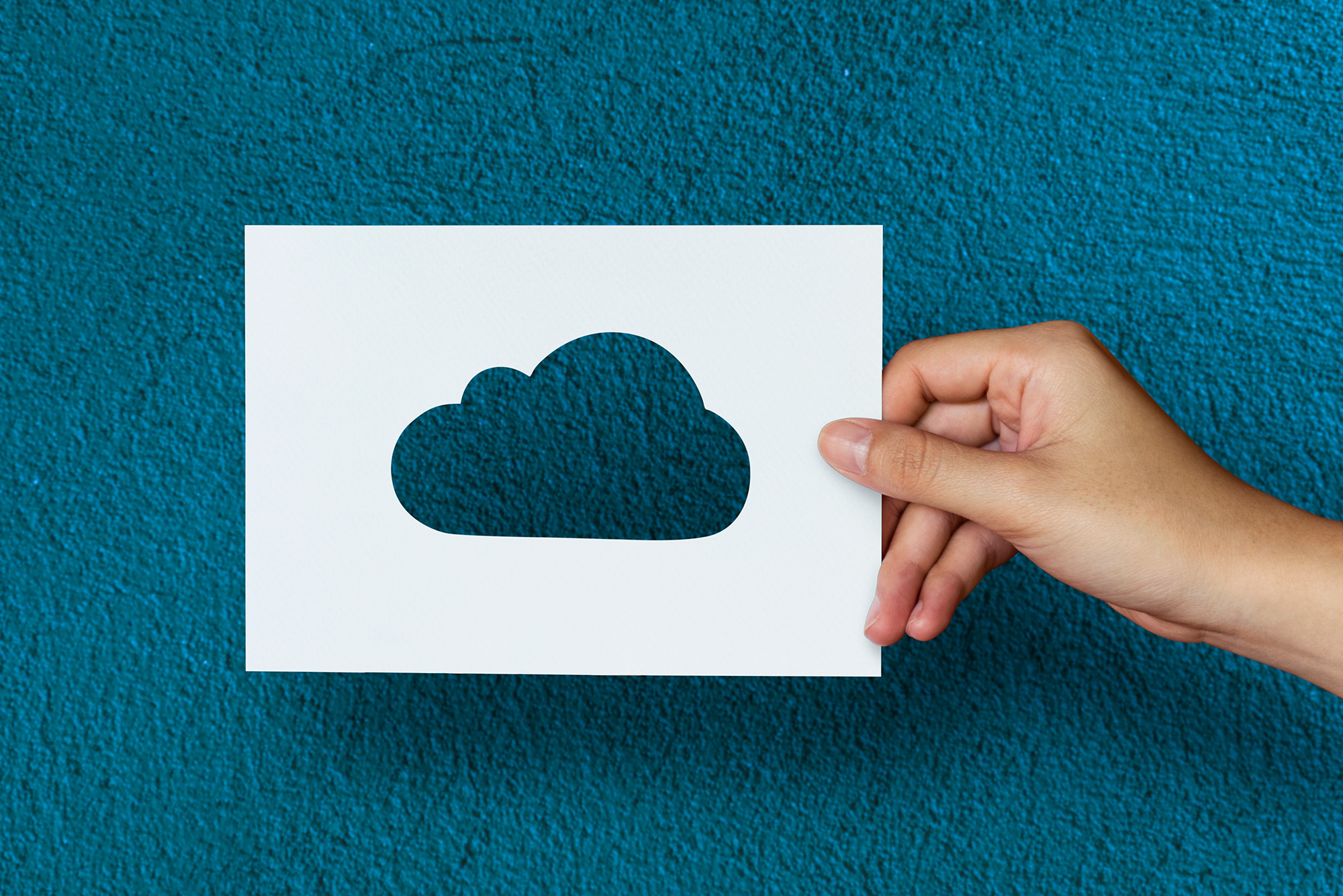 Hand holding a paper cutout of a cloud against a blue textured background, symbolizing cloud computing and digital storage.