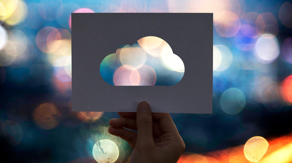 A person holding a paper cutout shaped like a cloud against a blurred city background with colorful lights, symbolizing cloud technology and digital transformation.