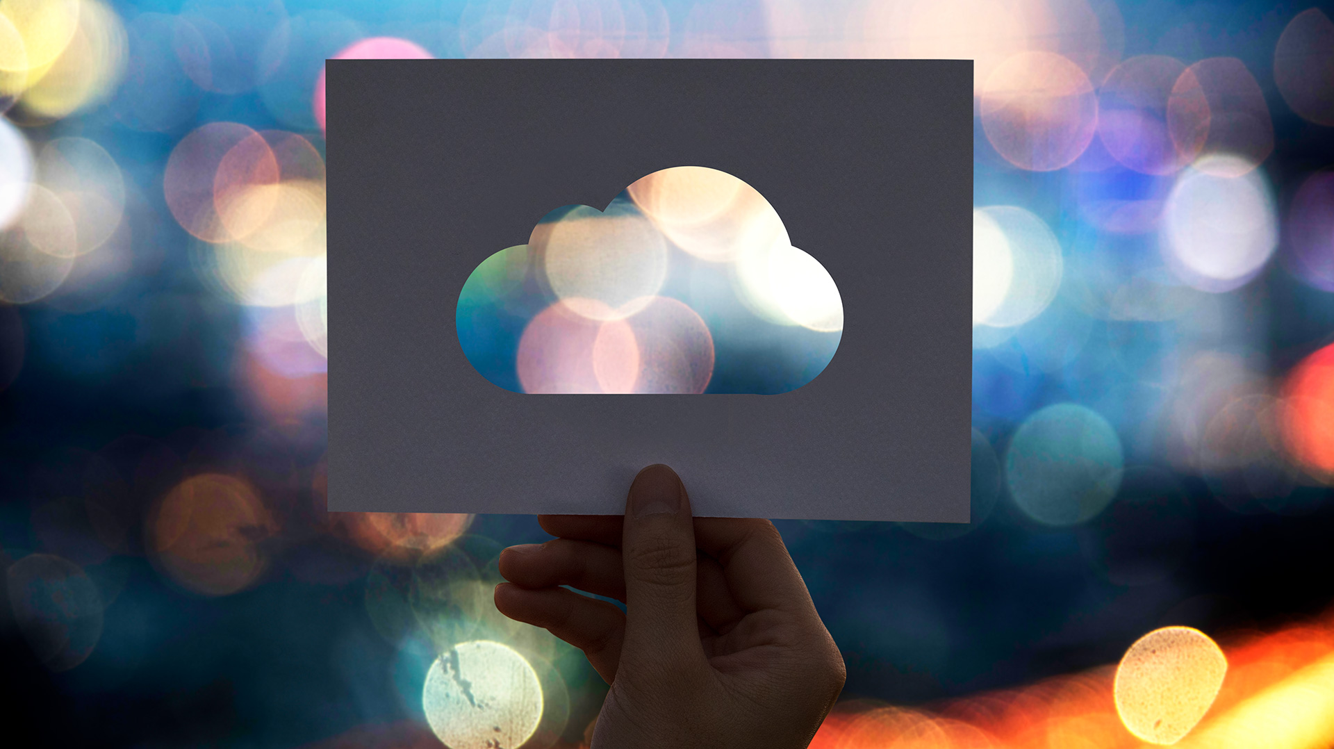 A person holding a paper cutout shaped like a cloud against a blurred city background with colorful lights, symbolizing cloud technology and digital transformation.