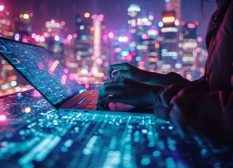 What is digital transformation A person types on a laptop with digital data visualizations projected across the keyboard and screen. The background features a vibrant, futuristic cityscape with colorful lights and a tech-forward atmosphere, symbolizing digital transformation and urban connectivity.